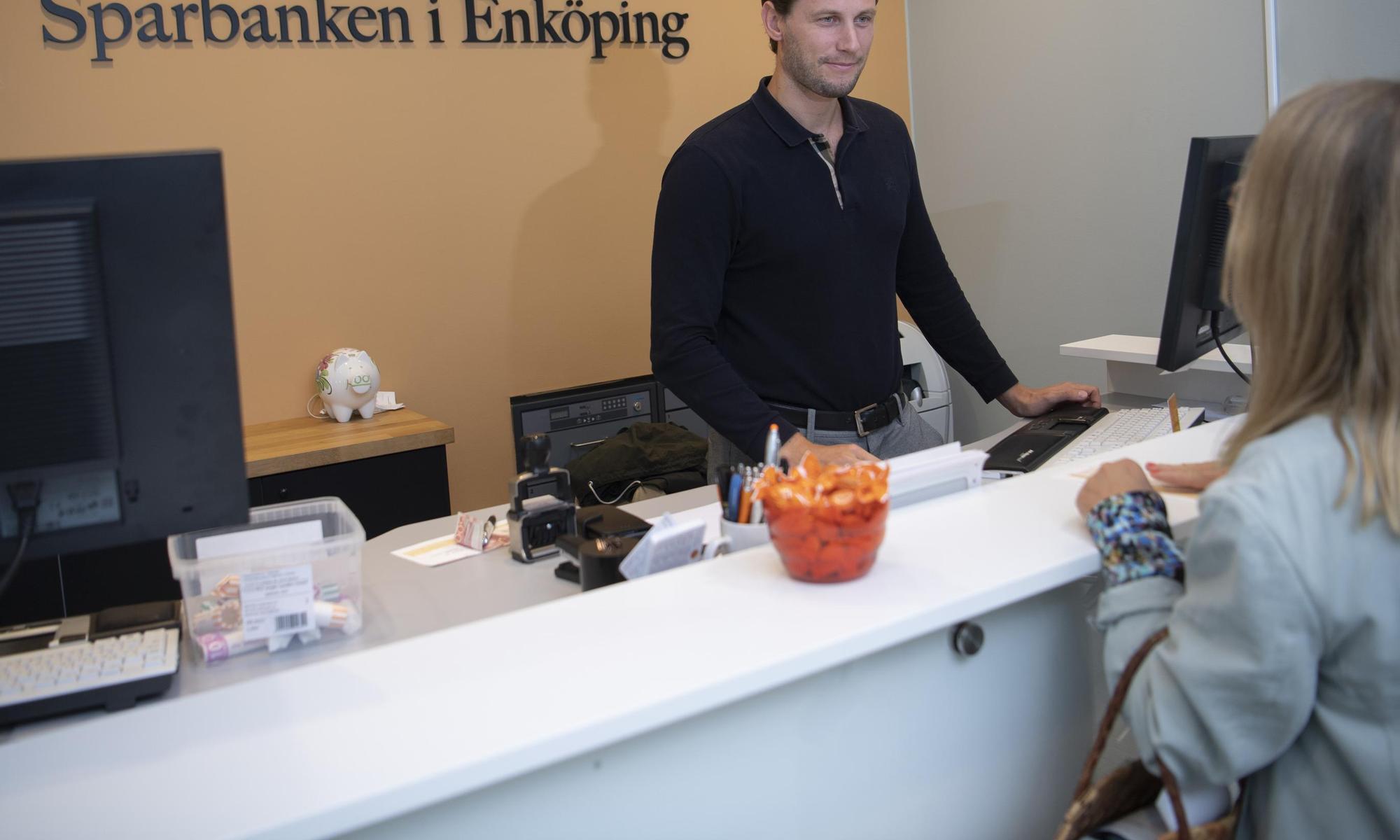 a man in a black shirt is standing next to a computer looking at a customer on the other side of the counter. Sparbanken i Enköping is written on the wall behind him.